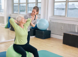 Mature woman working out on a fitness ball with help from personal trainer at gym.