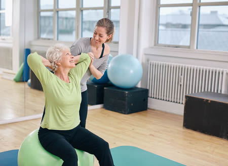 Mature woman working out on a fitness ball with help from personal trainer at gym. 